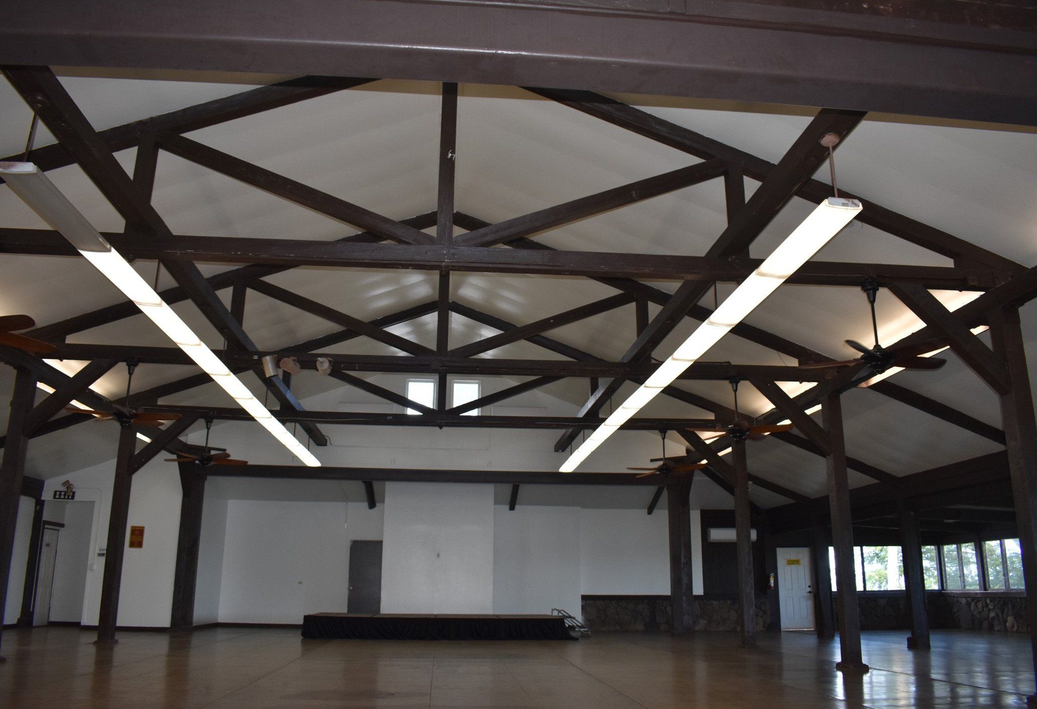 Hall at Heʻeia State Park with dark-stained timber roof trusses, a white ceiling and long light fixtures
