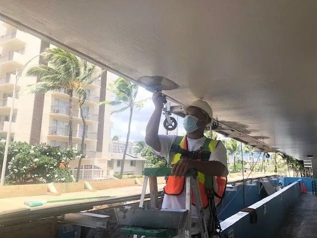 A painter in a hard hat and safety vest grinding a walkway ceiling at a high-rise