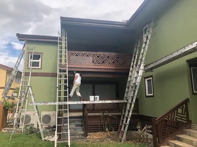 A painter on a plank between ladders, painting a green two-story house