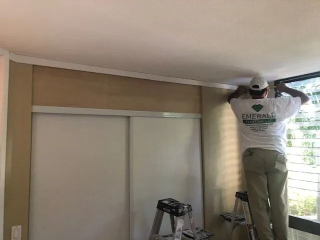 A painter on a ladder prepping the top of a bedroom wall beside a window